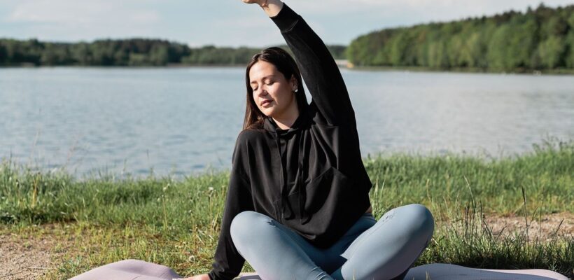 Kvinna sitter på yogamatta vid en sjö och sträcker armen över huvudet